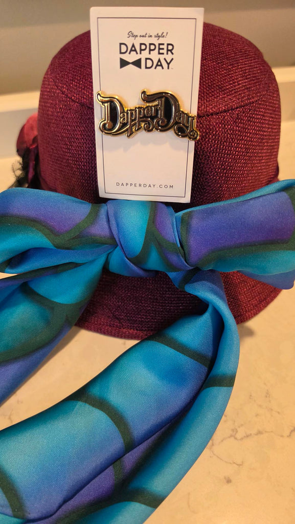purple cloche hat with mermaid scale scarf tied in a bow and a Dapper Day pin resting on top