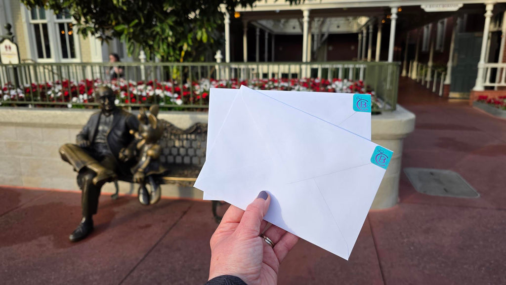 two letters with TheCB.Co mailing label on them being held up in front of the Roy E Disney and Minnie Mouse statue in Magic Kingdom
