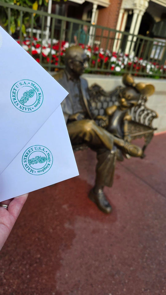 two letters with Main Steet U.S.A. seal stamped on the envelopes being held up in front of the Roy E Disney and Minnie Mouse statue in Magic Kingdom