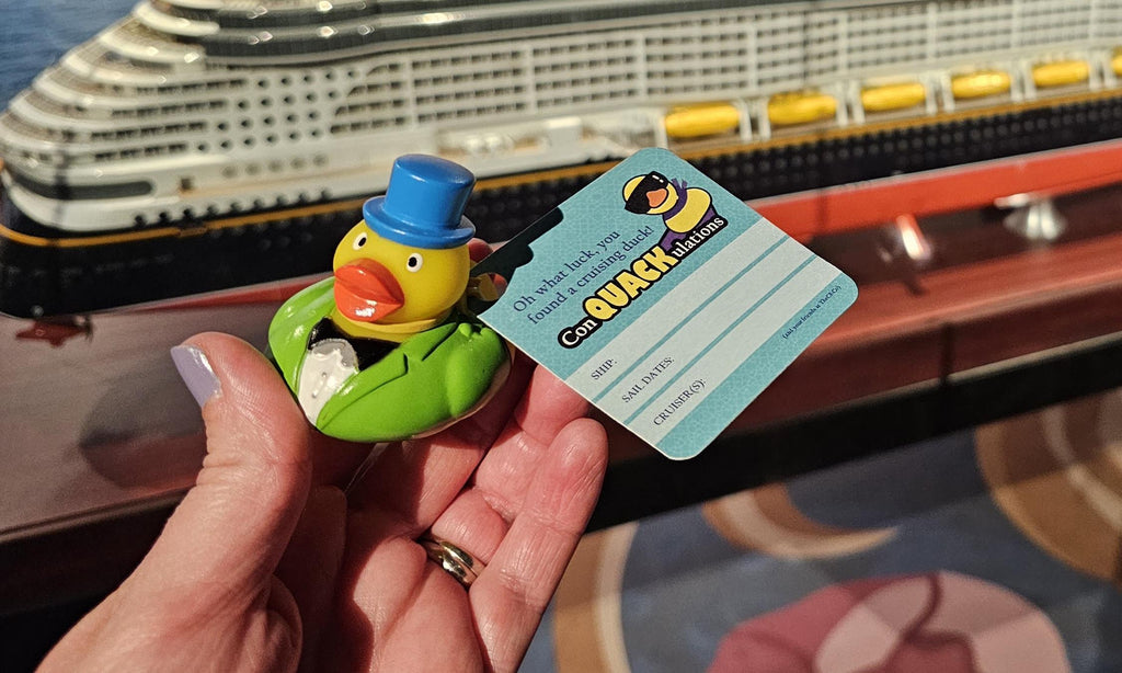 a rubber duck with a tag that says “ConQUCKulations” being held in front of a model of a cruise ship