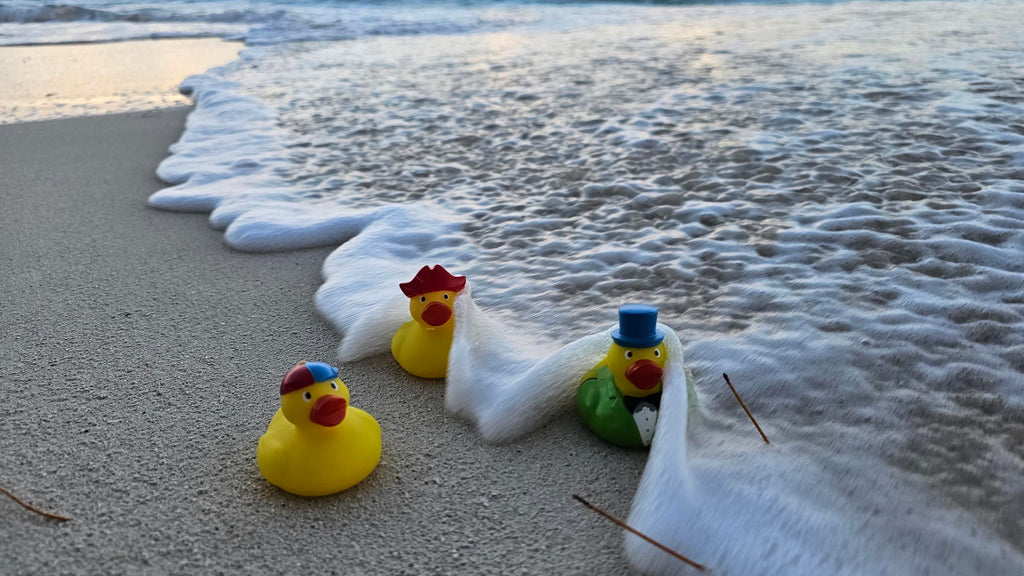 3 rubber ducks sitting on the beach with a wave crashing into them